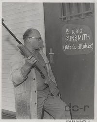 Schwarzweißfoto, Portraitaufnahme von Josef Fuchs mit einem Gewehr vor seiner Werkstatt